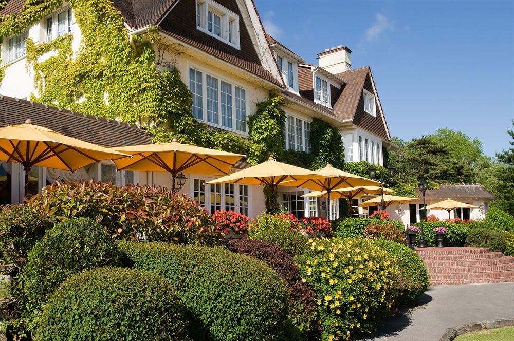 Le Manoir Hotel in the sunshine with garden bushes and parasols
