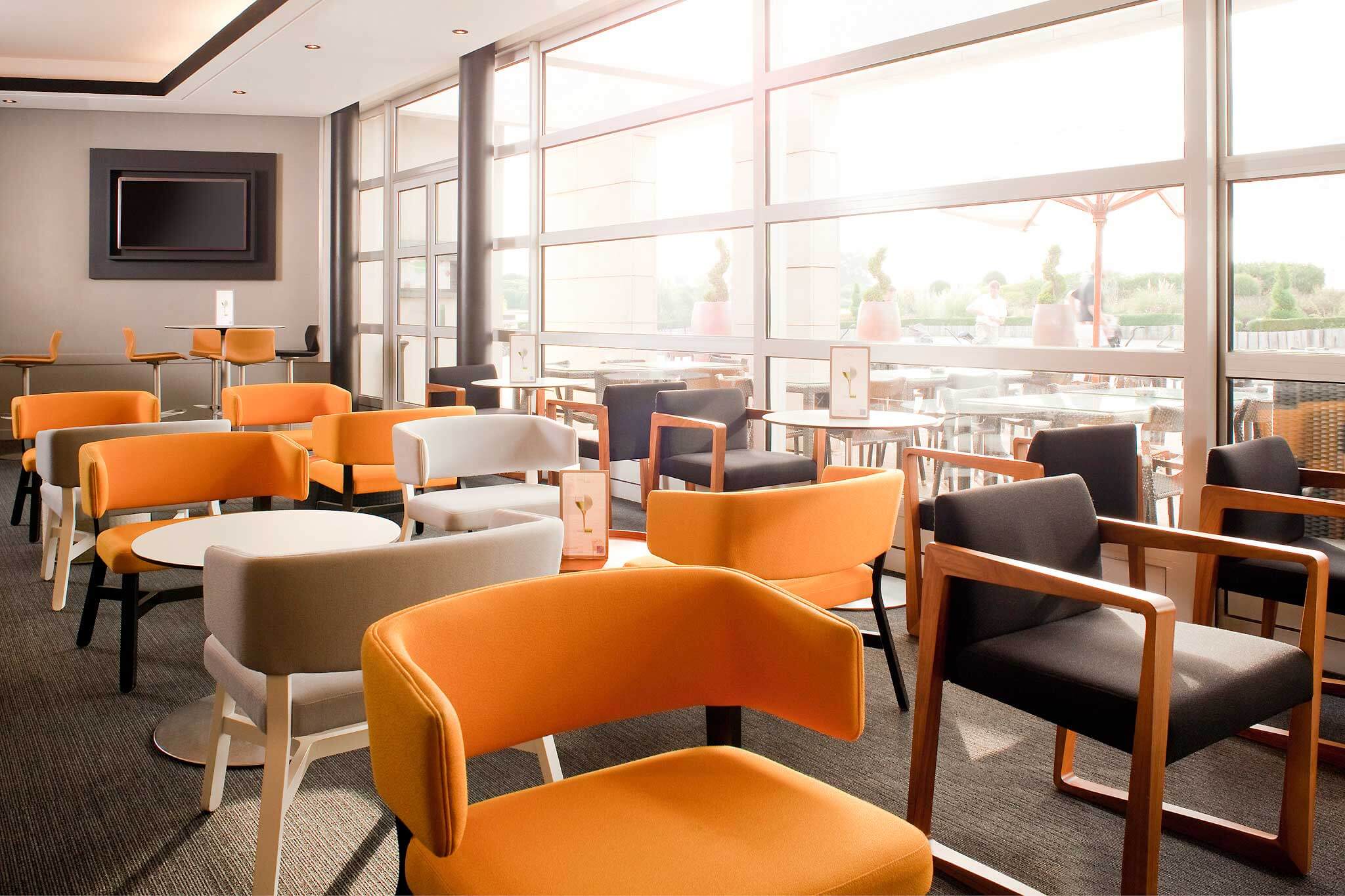 Novotel Saint Quentin Golf National's bar seating area with grey and orange chairs and tables and television on the wall