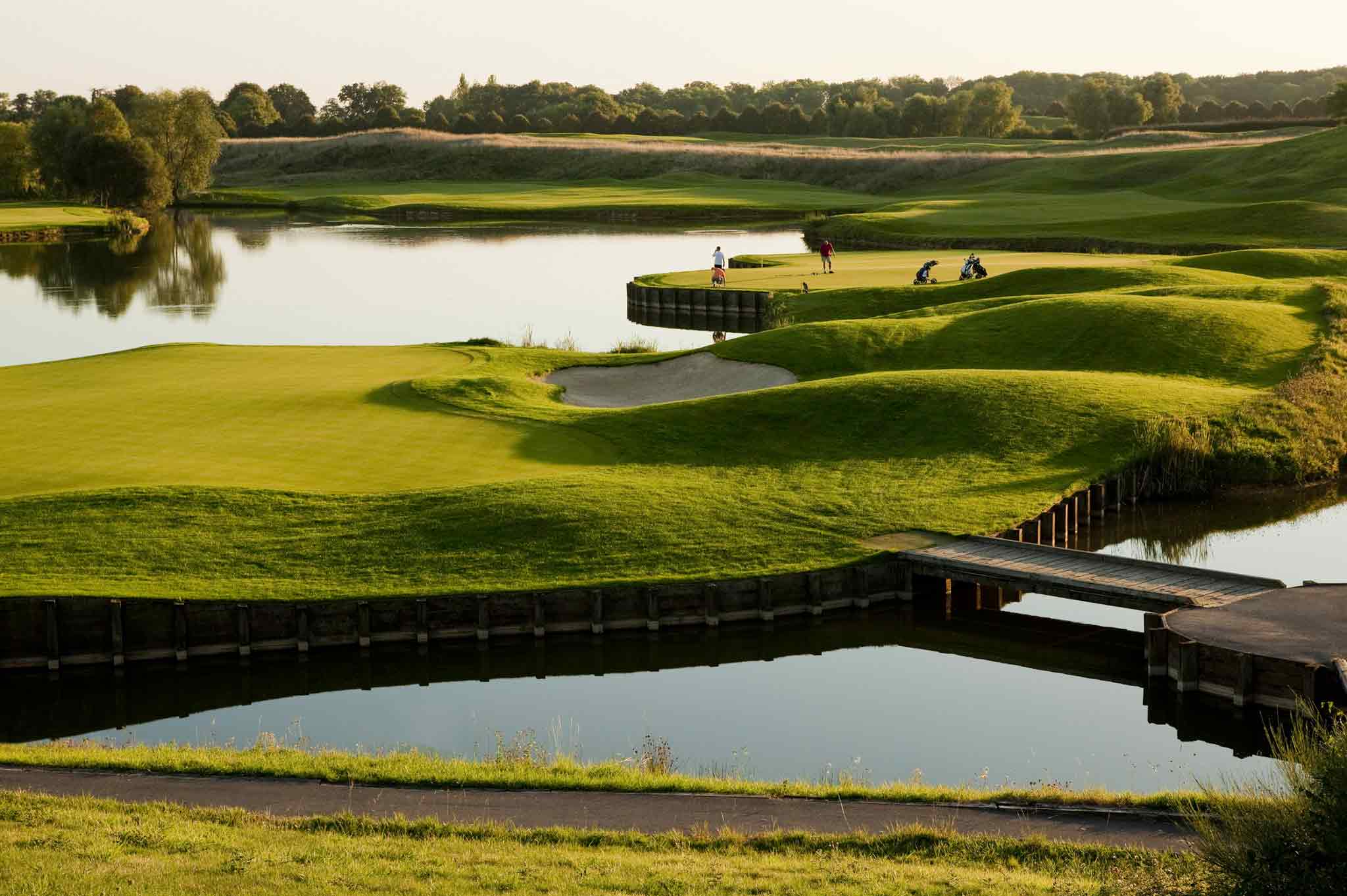 Two guests at Novotel Saint Quentin Golf National playing golf on the green with a lake and footbridge