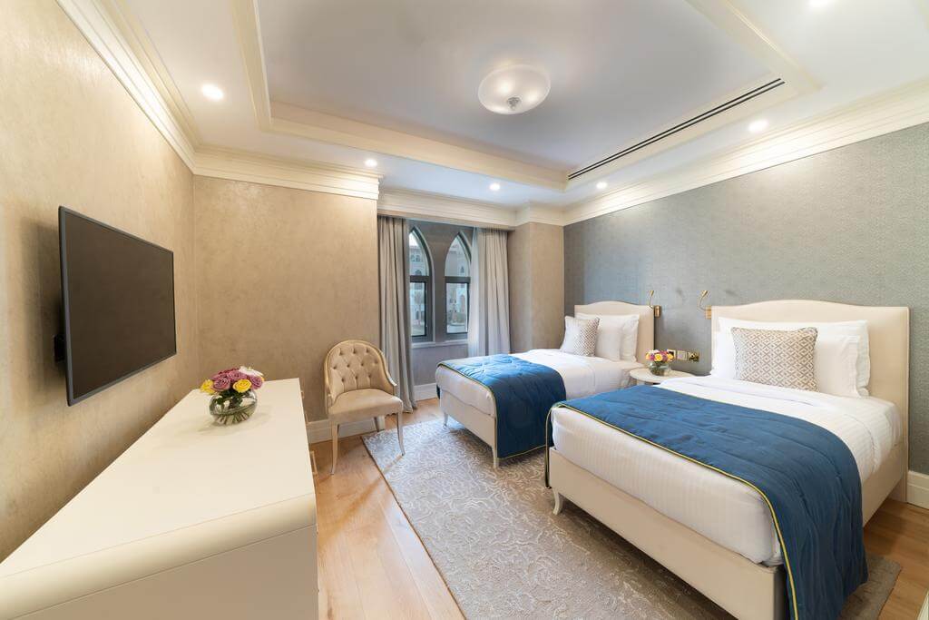 Twin bedroom at Rixos Saadiyat Island Abu Dhabi with flat screen television, decorative flowers on desk and side table, armchair, and blue throw on bed