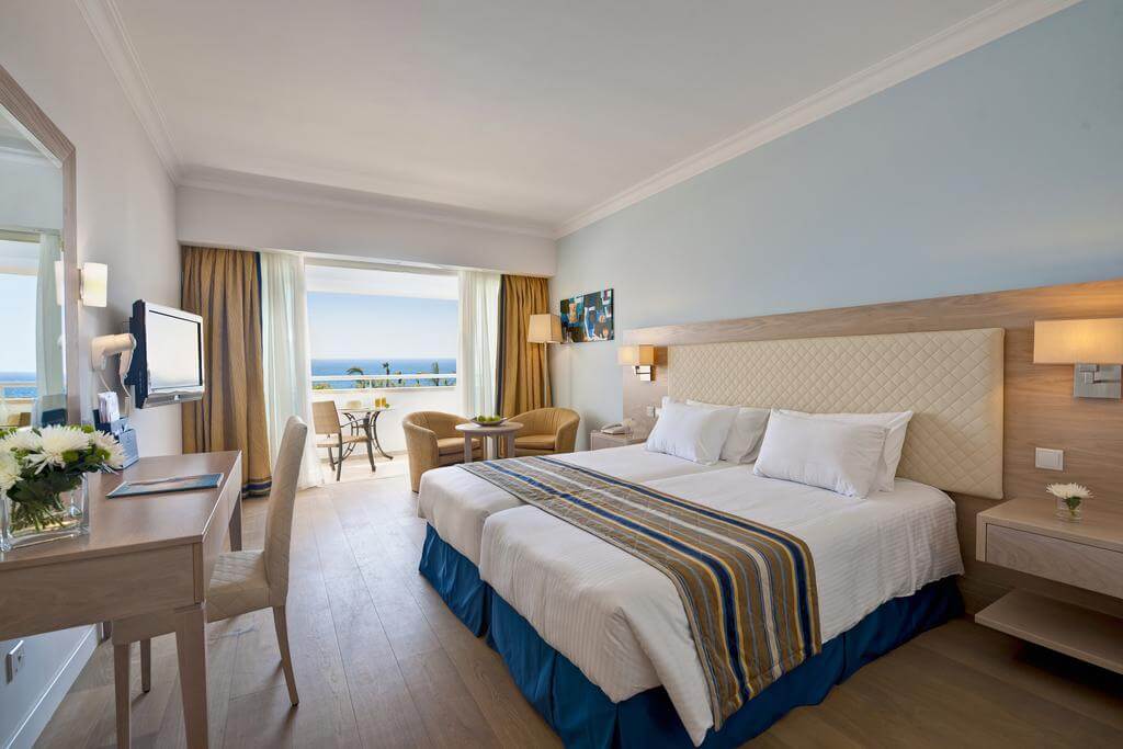 Twin bedroom at Olympic Lagoon Resort Paphos with television, desk, armchairs, white linen bedding and private balcony
