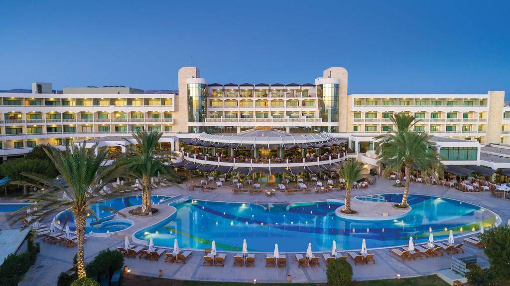 Exterior of Constantinou Bros Athena Beach Hotel overlooking the swimming pool, sun loungers and palm trees in the evening