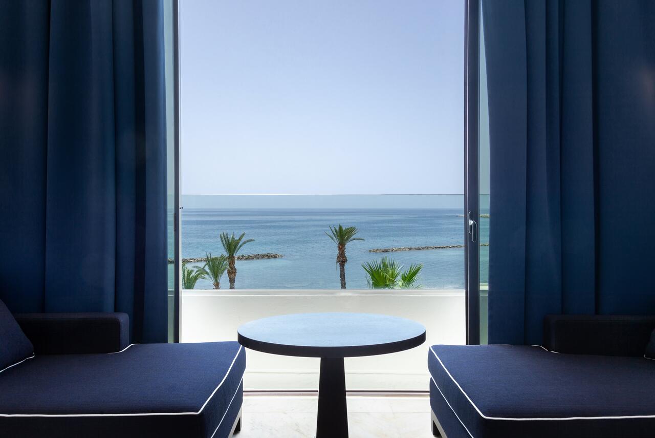 Blue chairs facing each other at Almyra Hotel with palm trees and blue sea through the patio doors